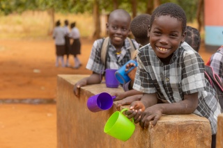 A boy leans over a wall, smiling with a mug in his hand, while his classmates stand behind him.